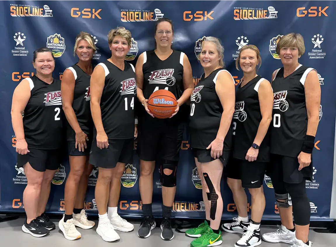 A women's basketball team poses for a picture in front of a branded backdrop.